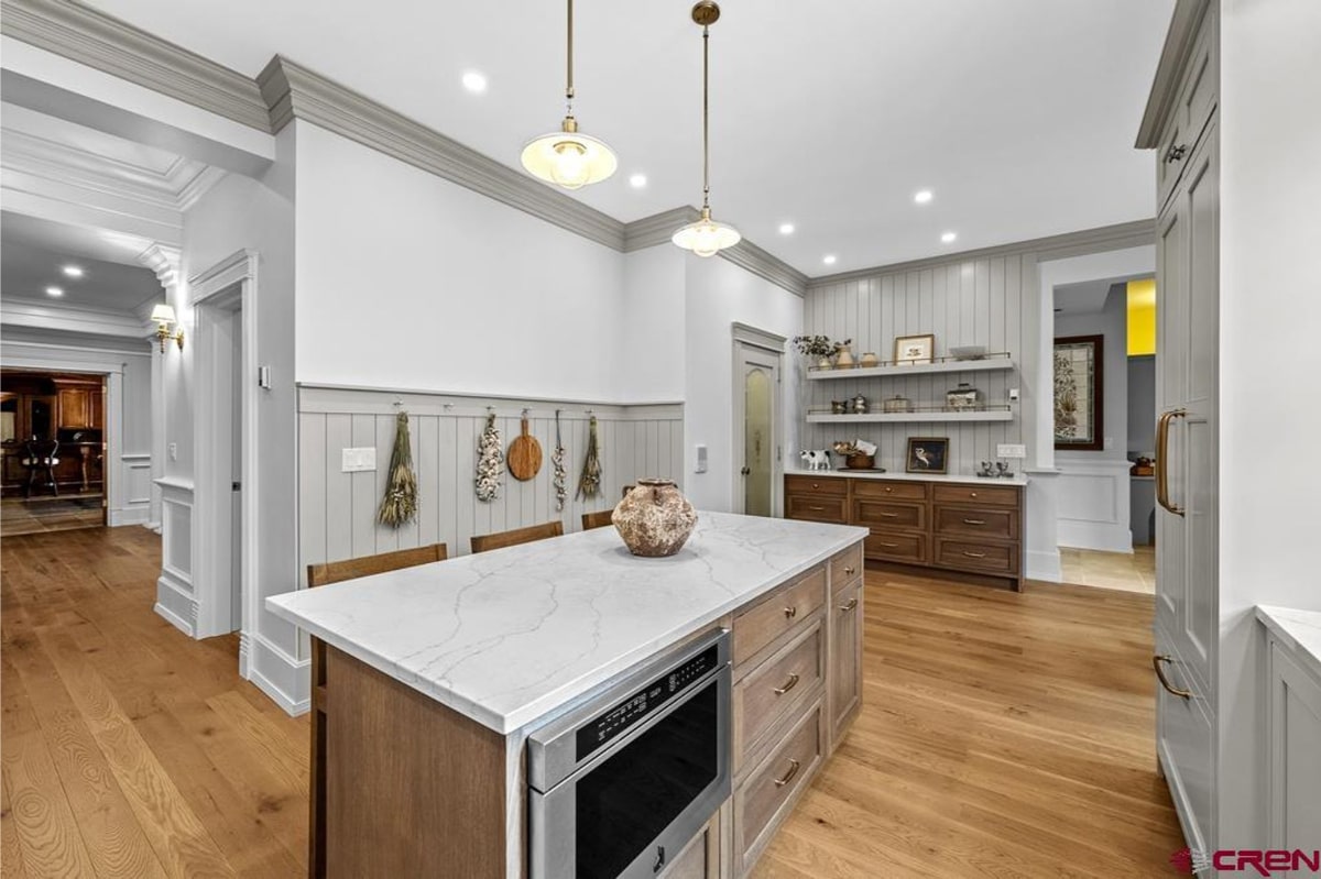 Kitchen with a large island in the center. The island has a white quartz countertop and light wood cabinets. There's a built-in microwave in the island. The walls have light gray shiplap paneling and open shelving. The floor is light-colored hardwood. The overall style is farmhouse or transitional.