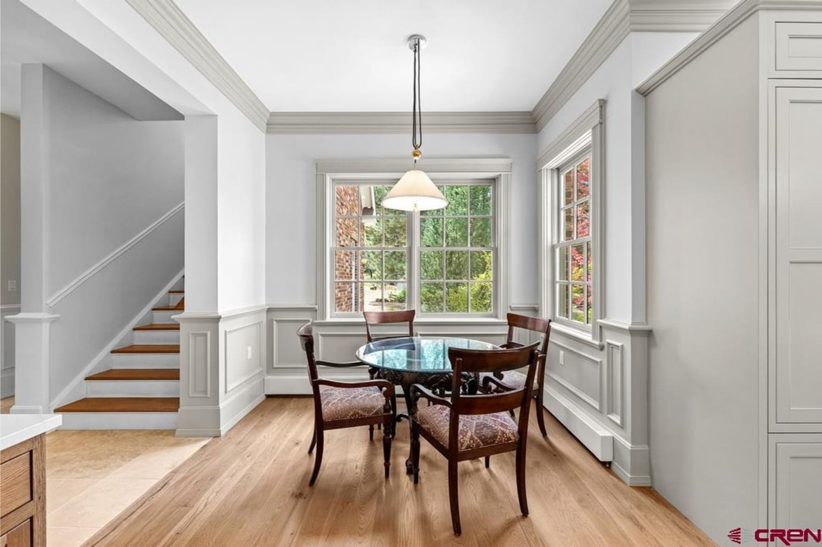 Dining room with light blue walls and white wainscoting. There's a glass-topped dining table with four dark wood chairs. Large windows provide natural light. A staircase is visible in the background. The overall style is traditional and elegant.