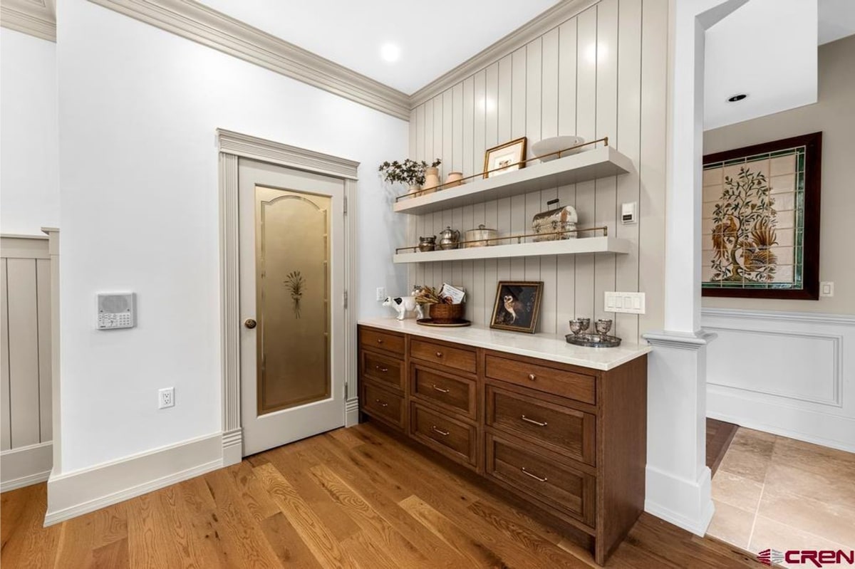 Kitchen pantry or butler's pantry with light gray shiplap walls. There are two floating shelves above a long set of drawers with a white quartz countertop. The style is farmhouse or transitional. A door with a frosted glass panel is visible to the left. The floor is light-colored hardwood.