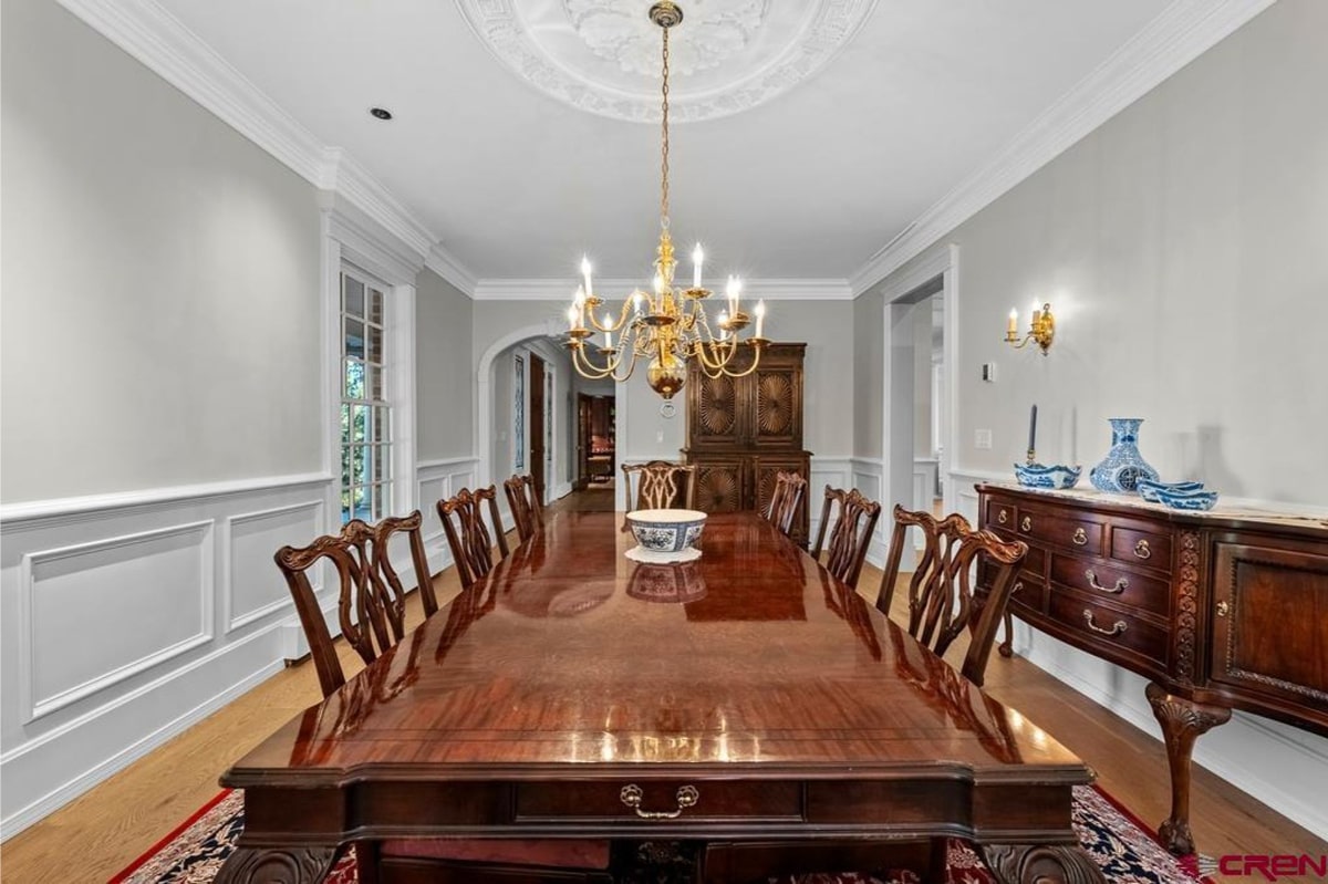 Formal dining room with light blue walls and white wainscoting. A large, highly polished mahogany dining table is set with chairs. A large chandelier hangs above the table. The overall style is traditional and elegant.