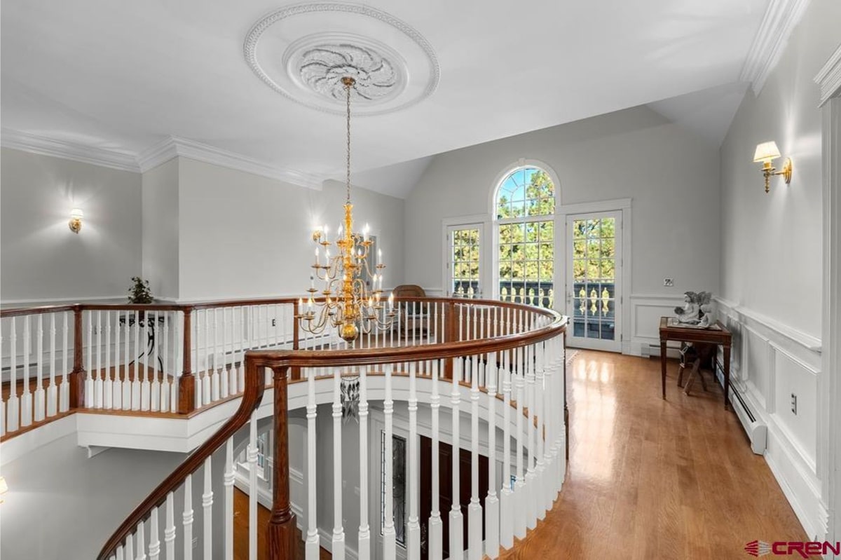 Curved staircase and upper landing with white railings and dark wood accents. A chandelier hangs above the landing. Large windows and French doors are visible in the background, offering a view of trees and greenery. The overall style is traditional and elegant.