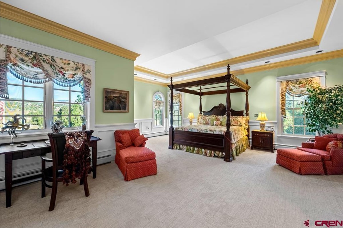 Master bedroom with a large, dark wood four-poster bed. The walls are a light green, and the floor is carpeted. There's a red chaise lounge and a small dark wood nightstand. Windows with curtains are visible, and a view of trees can be seen. The overall style is traditional and elegant.