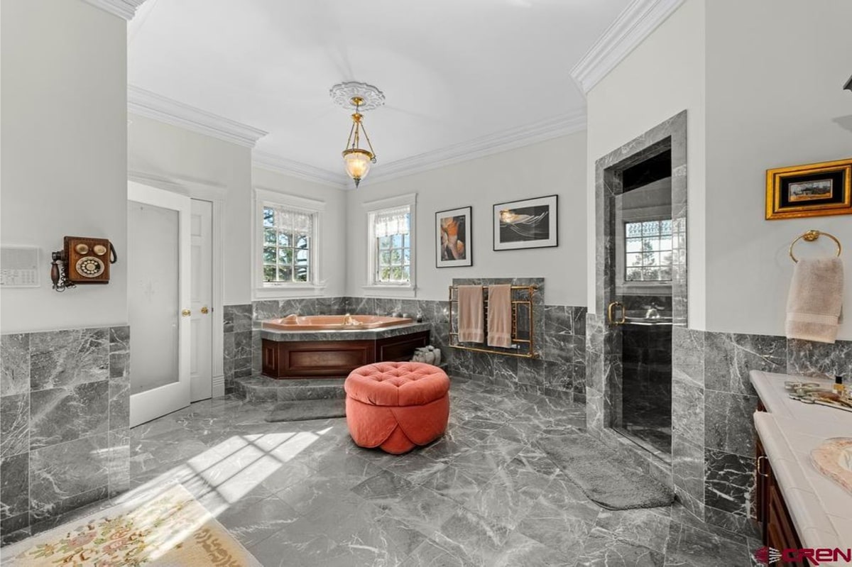 Luxurious bathroom with gray marble floors and walls. A large whirlpool tub is centered in the room. There's a pink ottoman and framed artwork on the wall. The overall style is elegant and spacious. Natural light streams in through windows.