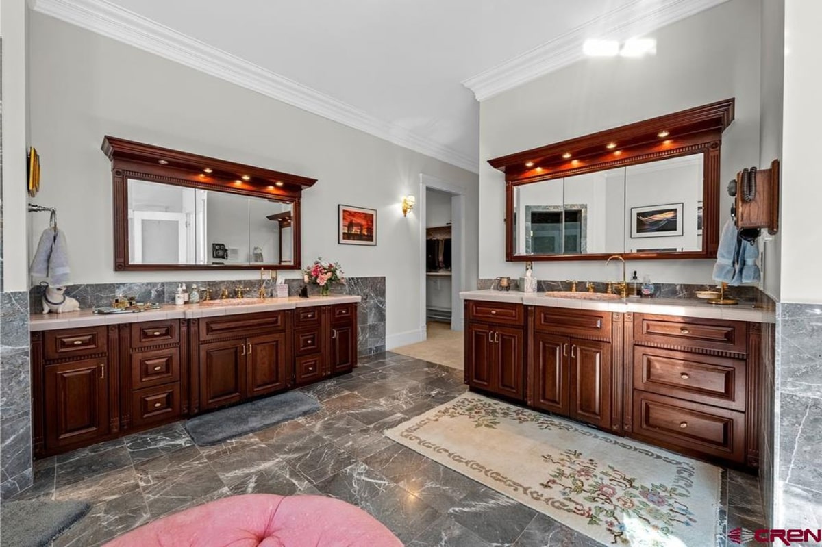 Large, luxurious bathroom with two double vanities. The vanities have dark wood cabinets and marble countertops. The floor is marble tile. The overall style is elegant and spacious. A rug is placed in front of the vanities.