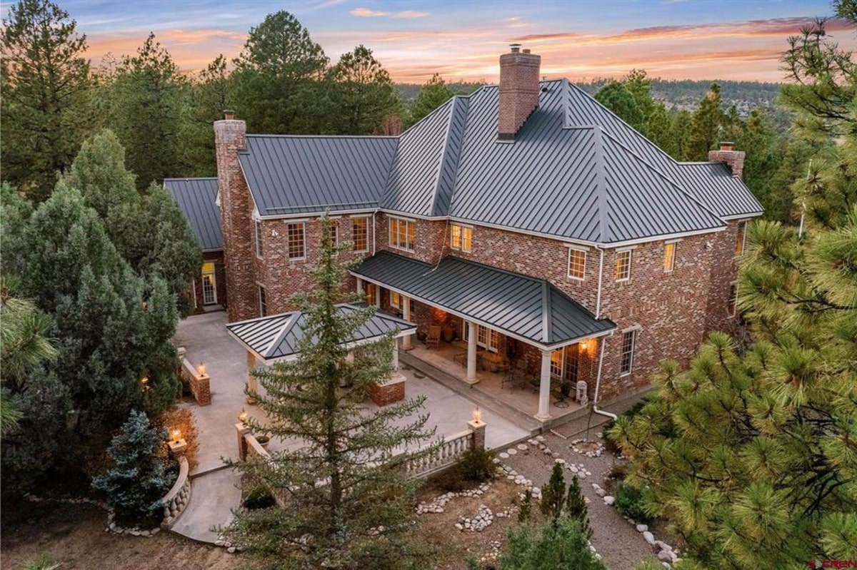 Elegant brick home features a large wraparound patio with detailed roofing and spacious outdoor areas. Warm interior lighting contrasts beautifully with the dark metal roof and lush greenery. Central courtyard design adds charm and enhances the house's inviting atmosphere.