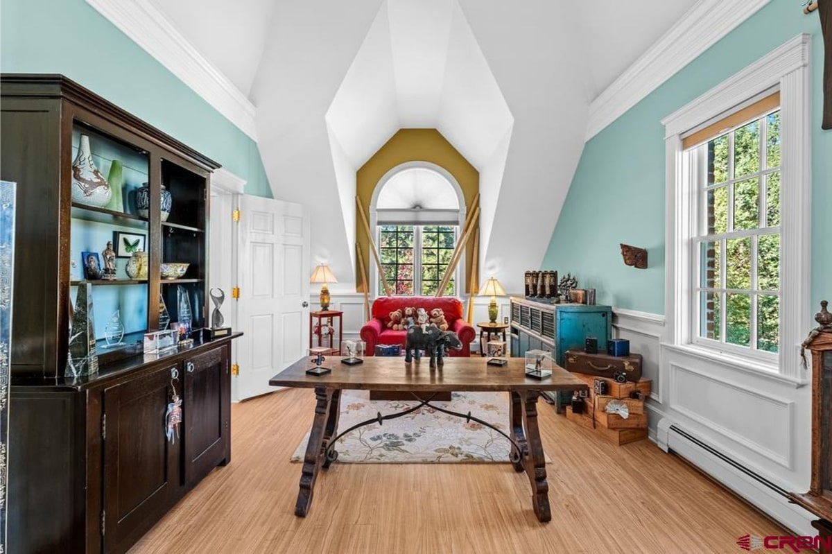 Room with vaulted ceilings and light teal walls. There's a large wooden table in the center of the room, and a red couch is visible in the background. There's also a dark wood cabinet and a teal cabinet. The overall style is eclectic and somewhat rustic. A large window provides natural light.