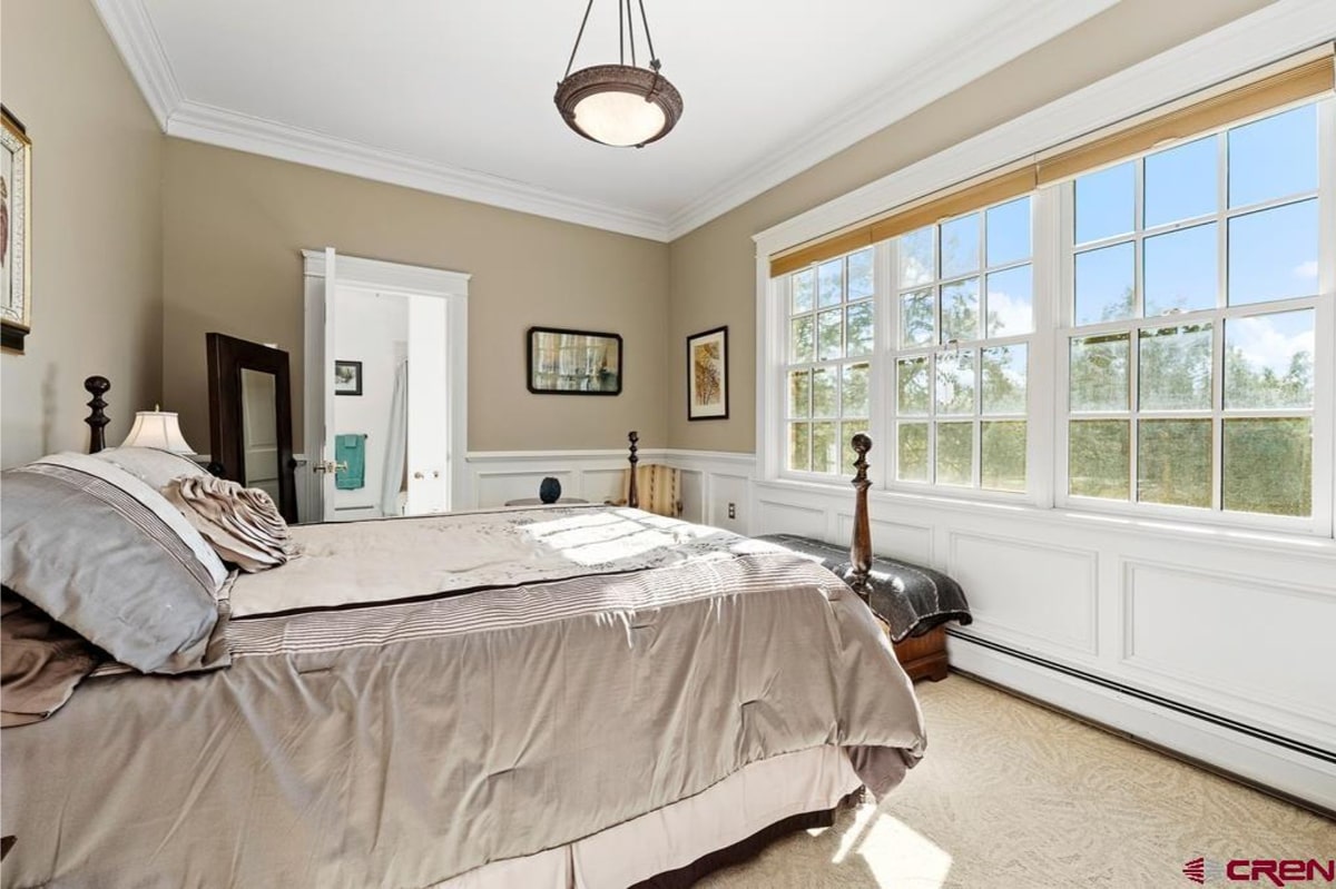 Bedroom with a large bed and a neutral color palette. The walls are a light beige, and the bed has a light gray comforter. Large windows provide natural light and a view of trees. The overall style is simple and elegant. There's white wainscoting on the lower portion of the walls.