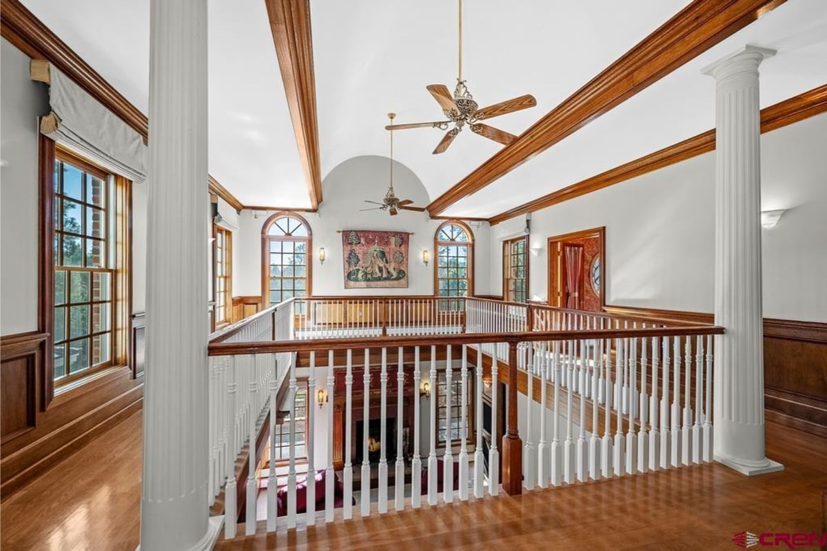 Hallway or balcony with white railings and dark wood beams. There are ceiling fans and windows. A tapestry is visible on the wall. The overall style is traditional and elegant. The view is from above, looking down.