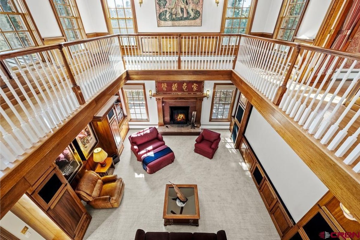 Great room or family room. The view is from above, looking down. There's a fireplace, seating area with leather and upholstered chairs and a sofa, and a coffee table. A balcony or second-story walkway surrounds the room. The overall style is traditional and somewhat formal. The floor appears to be light-colored stone or tile.