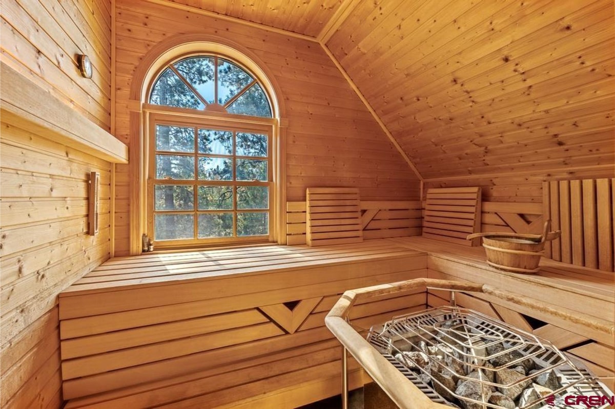 Sauna with wood paneling and a large arched window. There are wooden benches inside. A wood-burning stove or heater is visible. The overall style is rustic and natural. The window offers a view of trees.