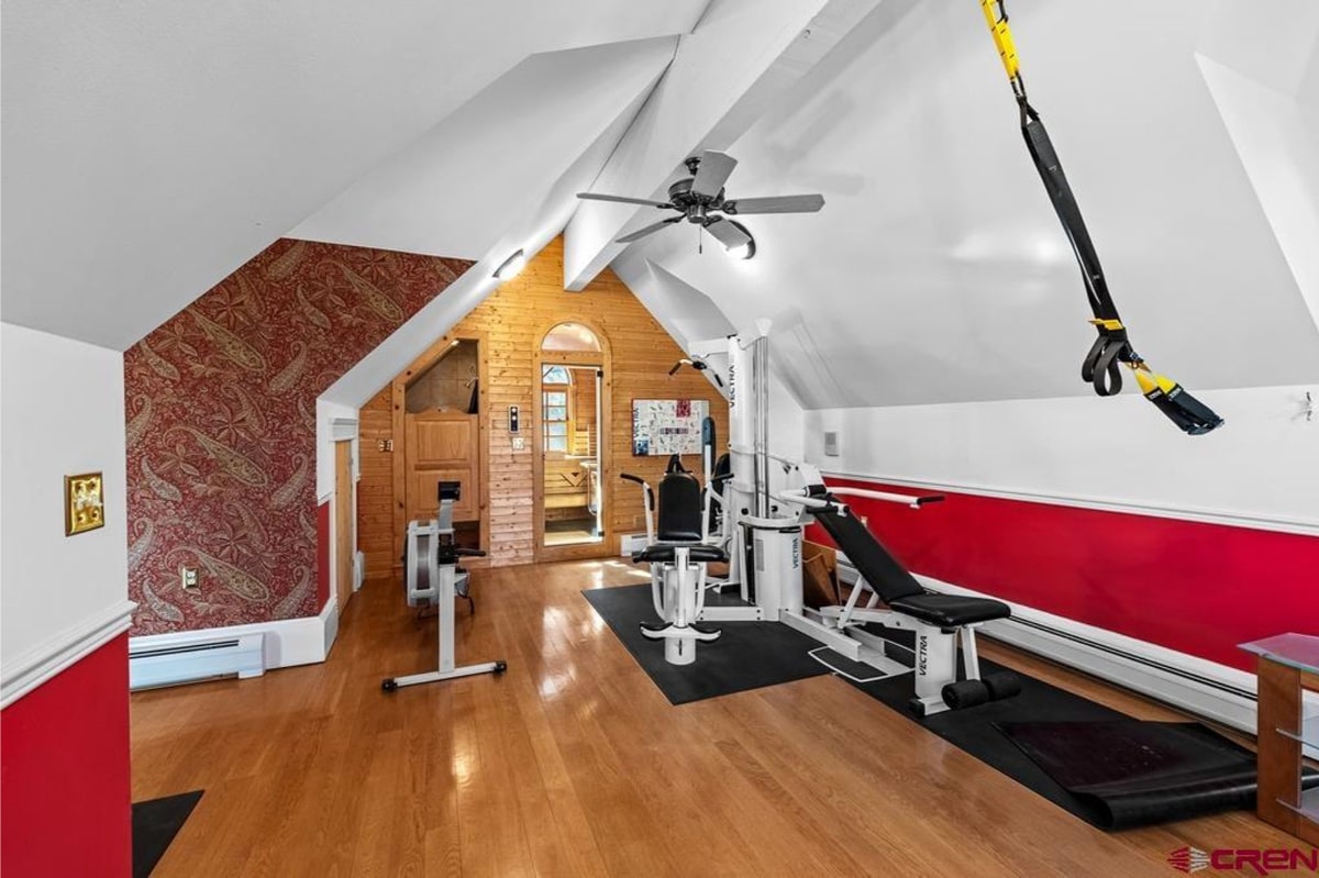 Home gym in an attic space. There's exercise equipment, including a weight machine and rowing machine. The walls are partially wood-paneled, and there's patterned wallpaper on one wall. The floor is hardwood. A ceiling fan is visible. The overall style is a mix of rustic and modern. There is a small door to another room.