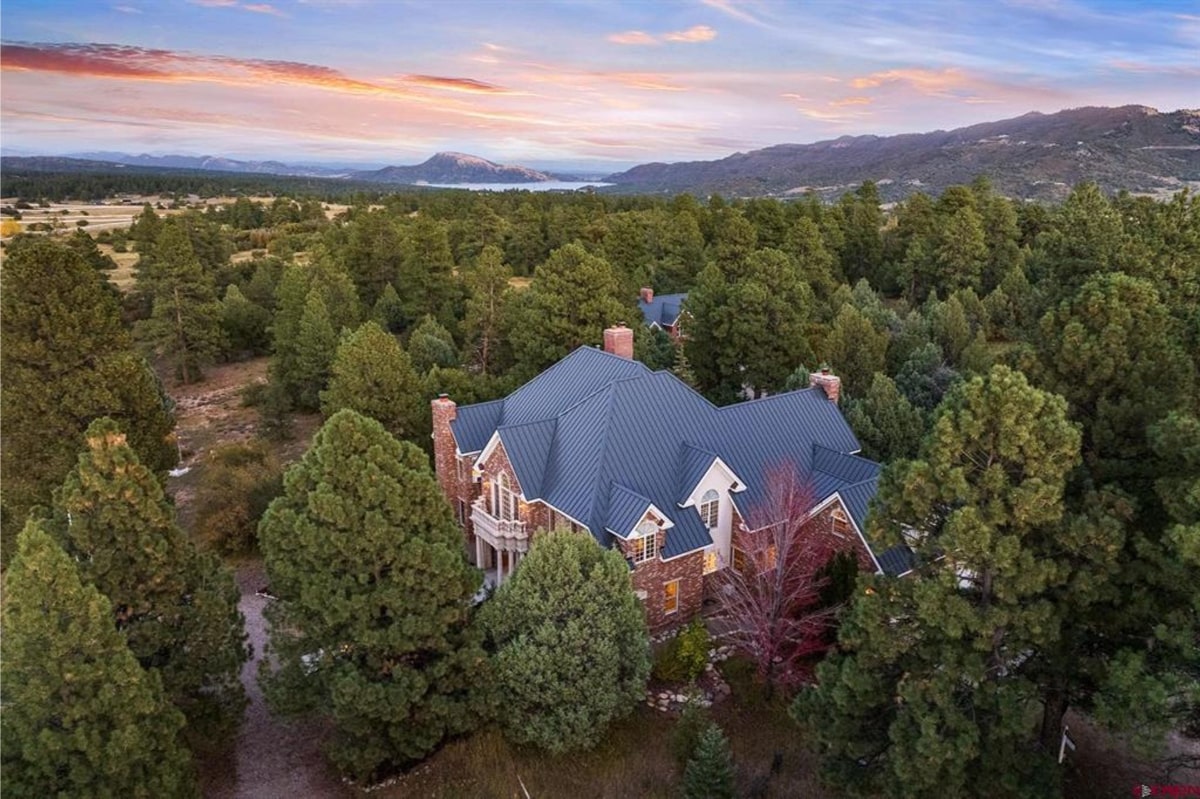 Aerial shot of a large, luxurious house with a teal metal roof. The house is made of stone and is surrounded by lush green trees.