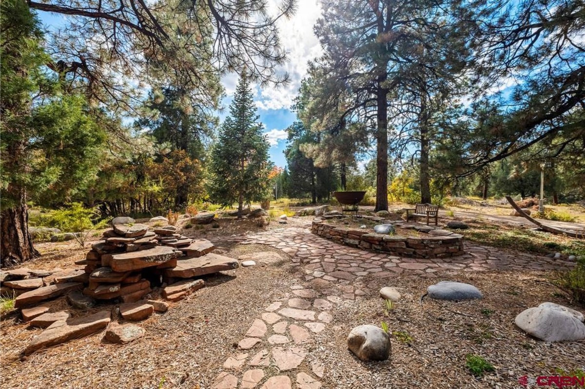 A stone pathway winds through a serene outdoor space, partially shaded by tall evergreen trees. A stone fire pit and seating area are visible, creating a tranquil and inviting atmosphere.