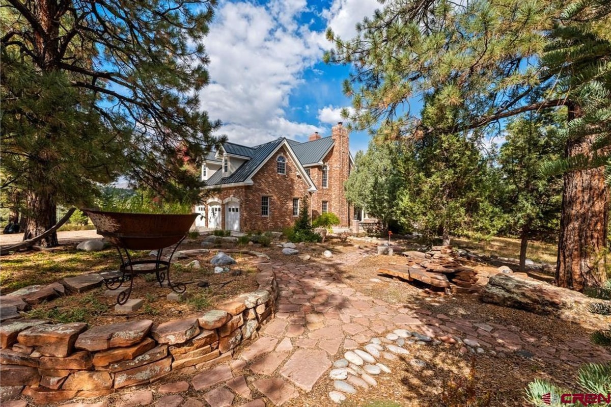 A brick house with a dark gray metal roof is partially visible through the branches of pine trees. A stone pathway leads to the house, which has a three-car garage. The scene is set against a bright blue sky with some clouds.