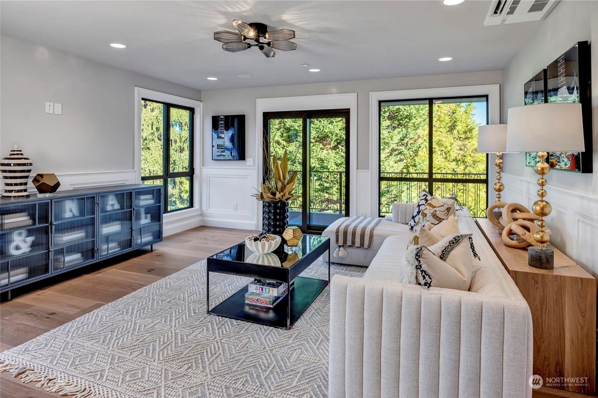 Living room with a white sofa, a coffee table, and a media. Large windows and sliding glass doors offer views of trees and a balcony. 