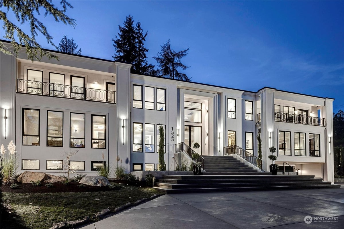 A modern, two-story white house with a grand staircase and large windows, illuminated at night.