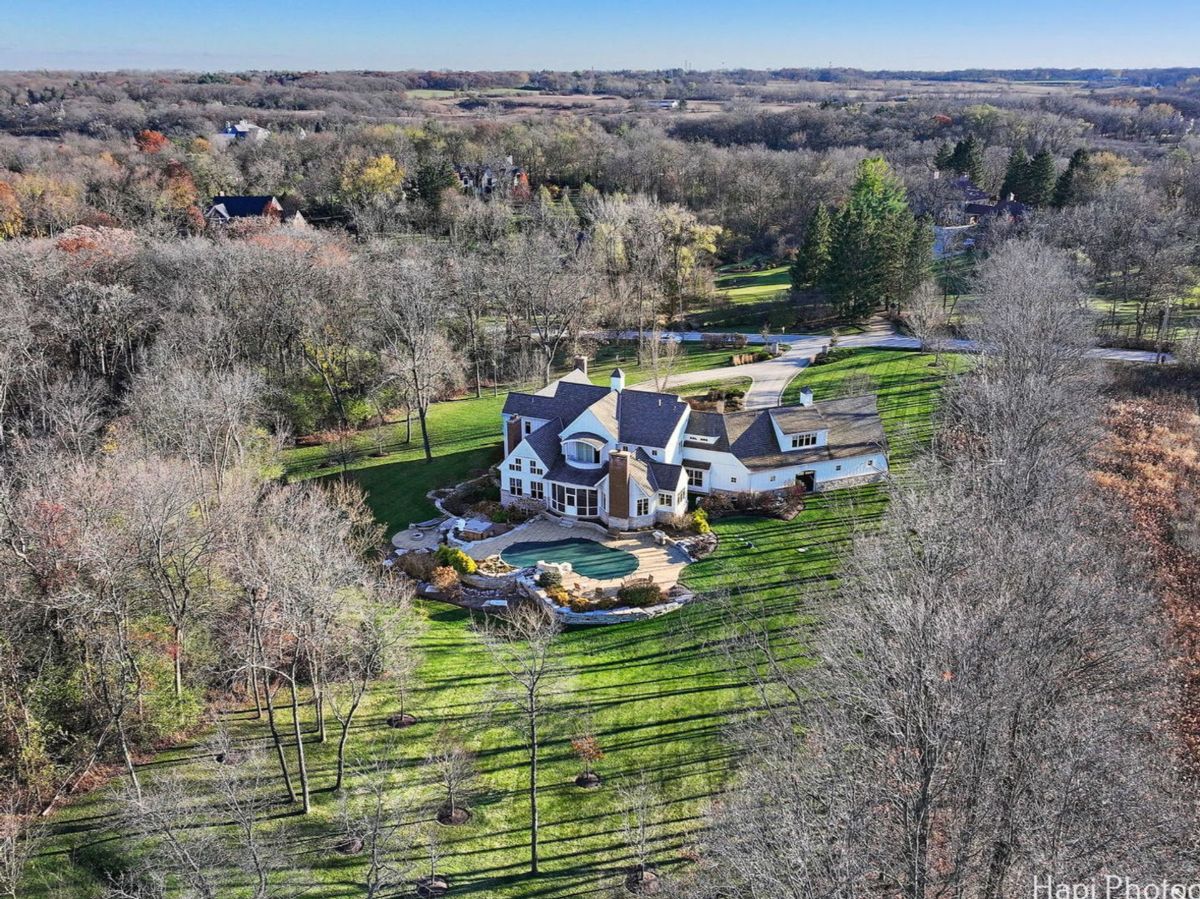 An aerial view showcases a sprawling white house nestled amidst a landscape of bare trees and a neatly manicured lawn, complete with a swimming pool.