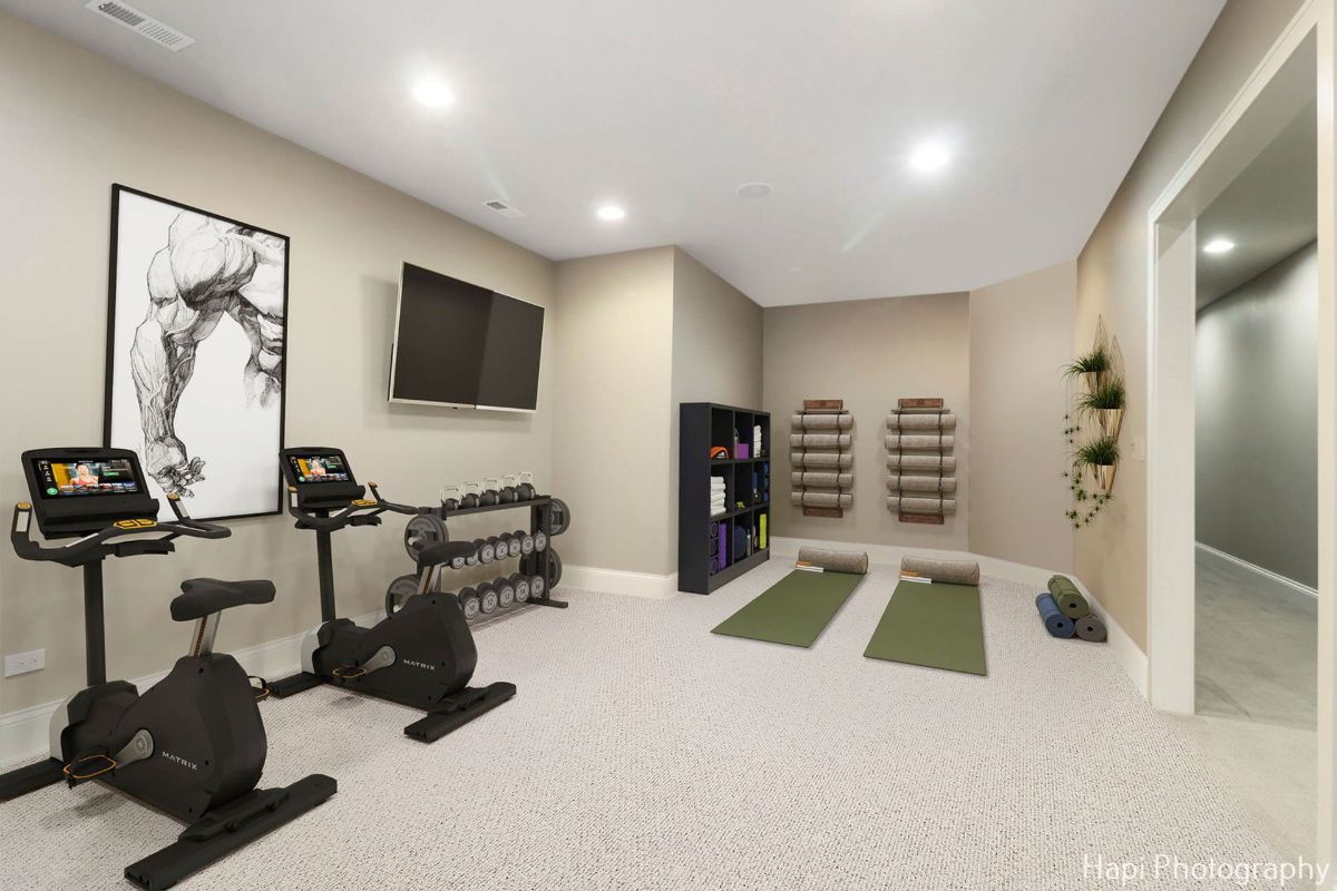 Home gym with two stationary bikes, a dumbbell rack, yoga mats, and a television provides a complete fitness space.