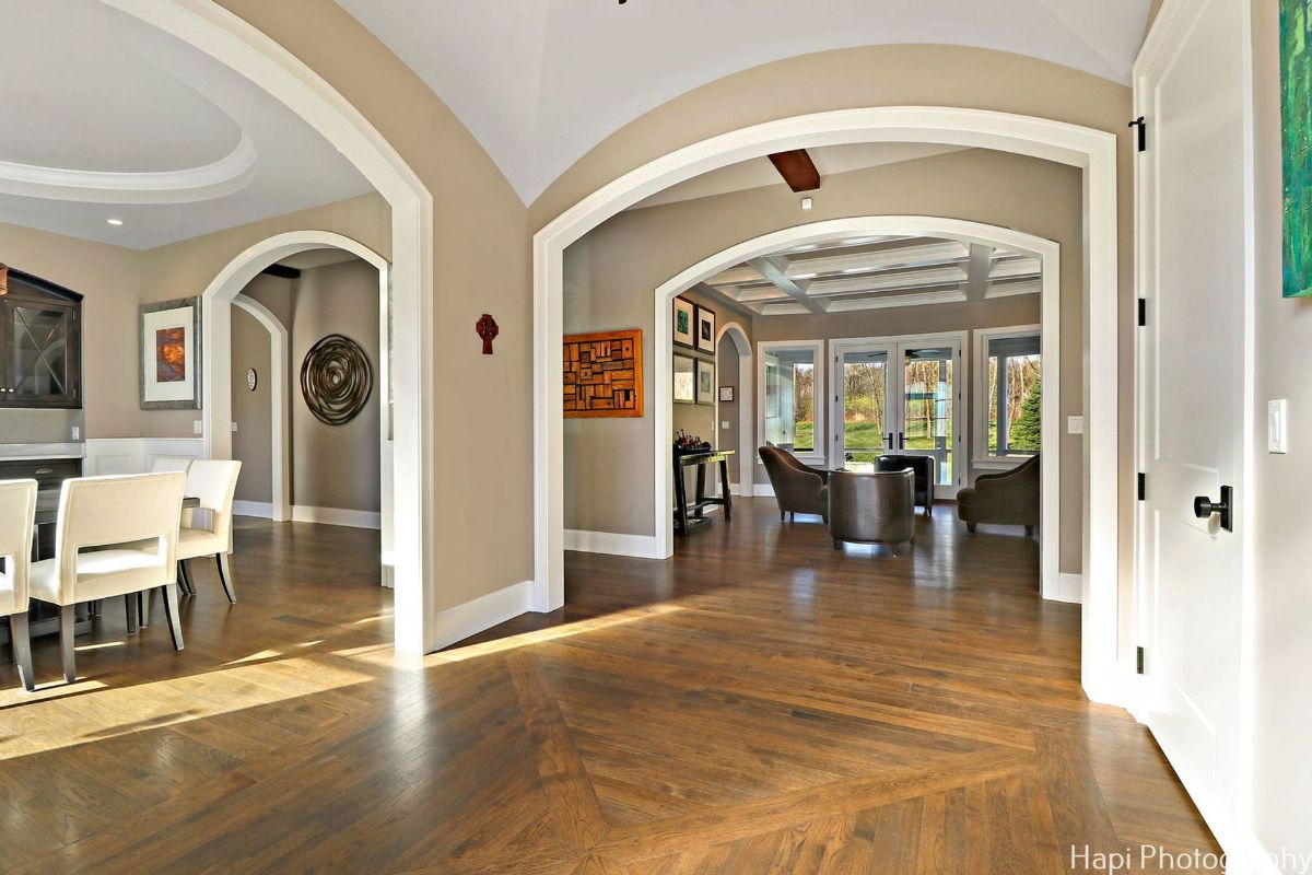 Open-plan interior with hardwood floors laid in a herringbone pattern. Arched doorways lead to a dining area and a sitting room with large windows overlooking a verdant outdoor space.