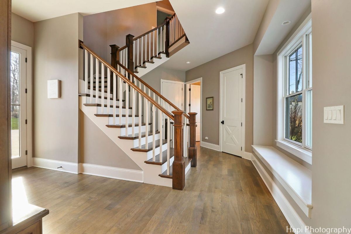 Light-colored hardwood floor hallway with a staircase featuring a dark wood handrail and white balusters.