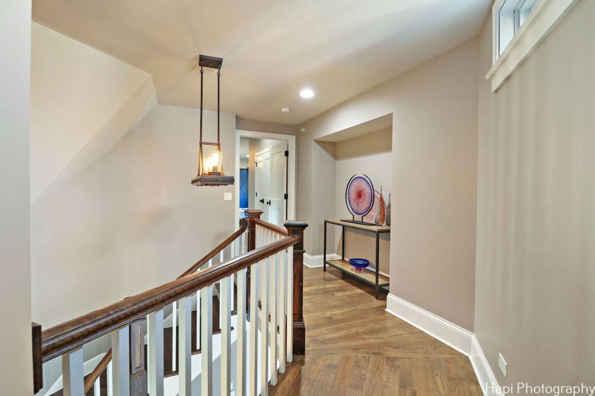 Hallway with light gray walls, hardwood floors, a wooden staircase with white spindles, and a modern light fixture hanging from the ceiling.