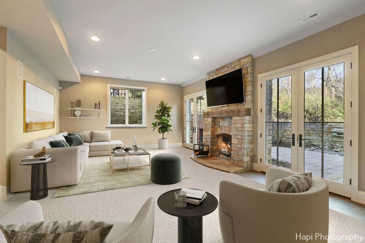 Luxurious basement family room with a stone fireplace, large windows offering an outdoor view, and a light beige sectional sofa.