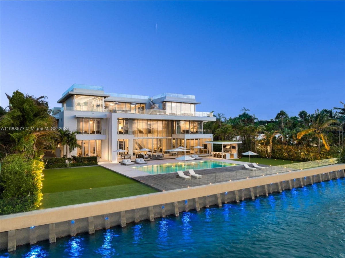 Mansion has extensive glass windows and is situated on a grassy lawn next to a swimming pool. A seawall separates the property from the water.