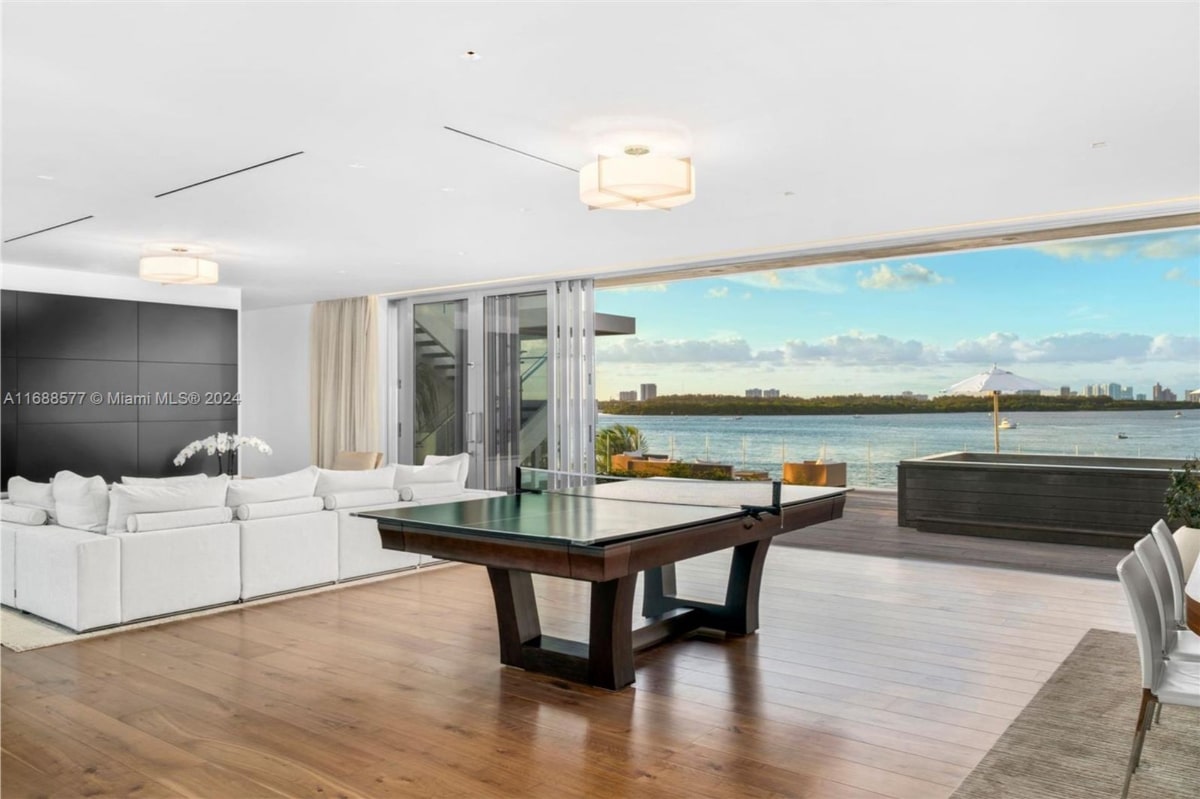 Game room with dark-wood table tennis table is the focal point, situated in a room with hardwood floors and large windows offering a stunning waterfront view. White couches are visible in the background.