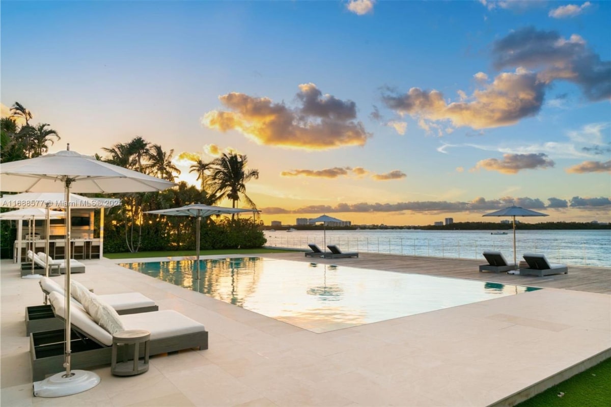 Tranquil poolside scene at sunset. A large, rectangular pool is the central feature, surrounded by lounge chairs and umbrellas. Palm trees and a waterfront are visible in the background.