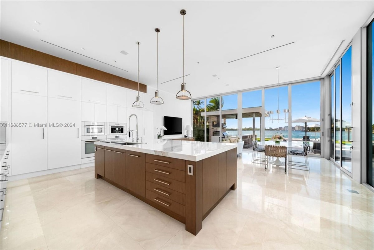 Kitchen with a large island and white cabinetry. The kitchen has a bright, open feel with floor-to-ceiling windows offering a stunning view of a waterfront patio and beyond.