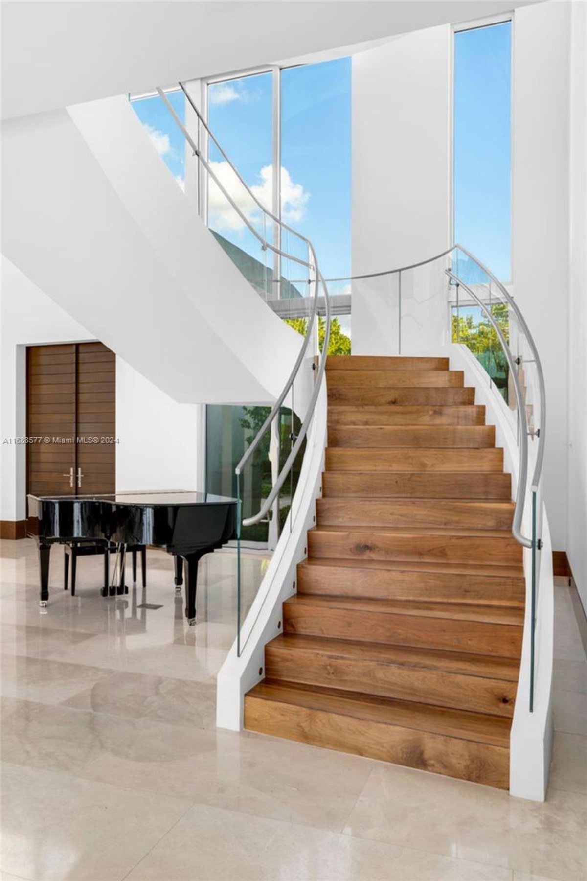 Grand staircase with dark wood steps and a modern, curved metal railing. A black grand piano is situated at the base of the stairs in a spacious area with polished floors.