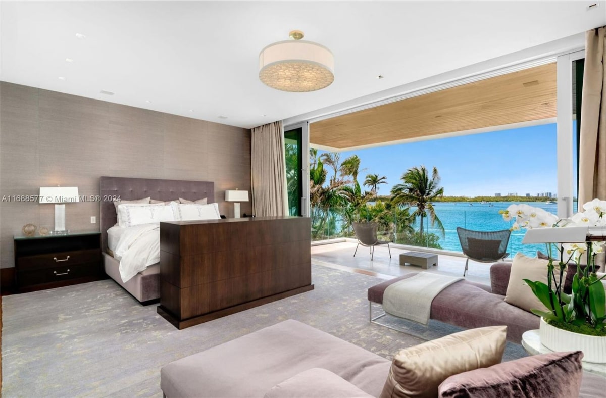  Bedroom with a view of a tropical waterfront. The bedroom features a large bed with a dark wood media cabinet at its foot, and floor-to-ceiling windows open to a private balcony overlooking a stunning ocean view with palm trees.