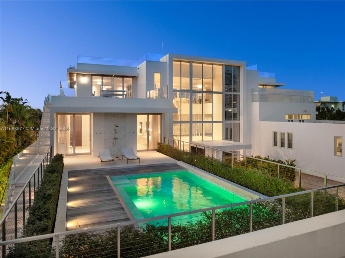 Nighttime view of a modern home with a rooftop pool. Landscaping is visible around the pool and the house.