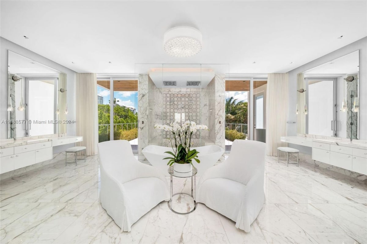  Bathroom with a spa-like atmosphere. The bathroom features a large soaking tub, white marble floors and walls, and two white lounge chairs. Large windows provide a view of lush greenery and a hint of a waterfront.