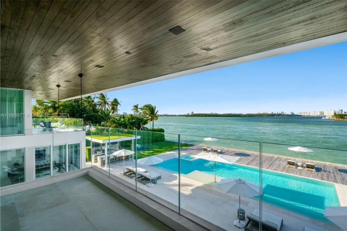 View from a balcony overlooking a large, modern pool and a waterfront. The pool is long and rectangular, and there are lounge chairs and umbrellas around it. Beyond the pool, there's a wide expanse of water and a distant shoreline.