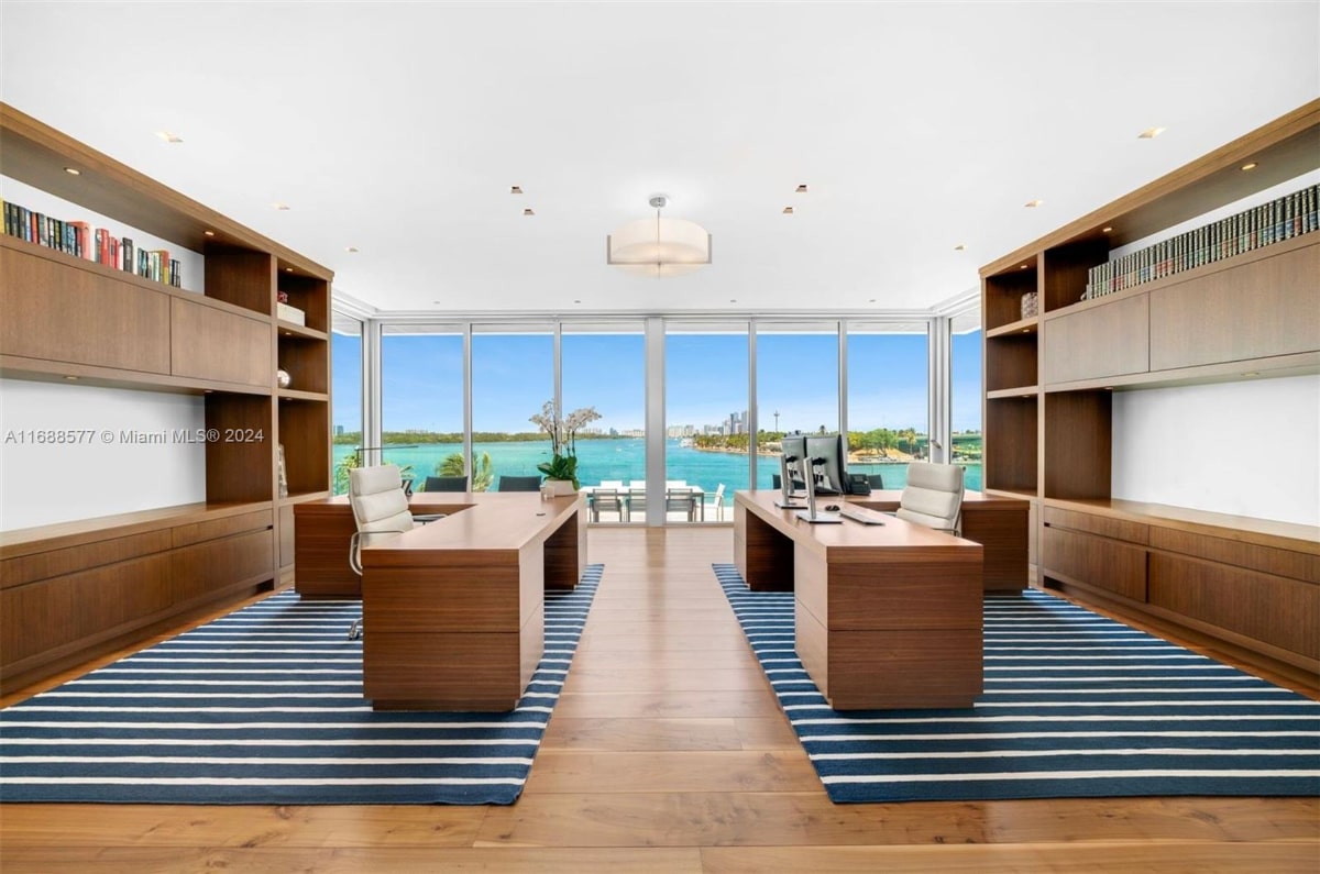 Home office with two large desks facing each other. The office boasts floor-to-ceiling windows offering a stunning waterfront view. Built-in wooden cabinetry and a blue and white striped rug complete the sophisticated design.