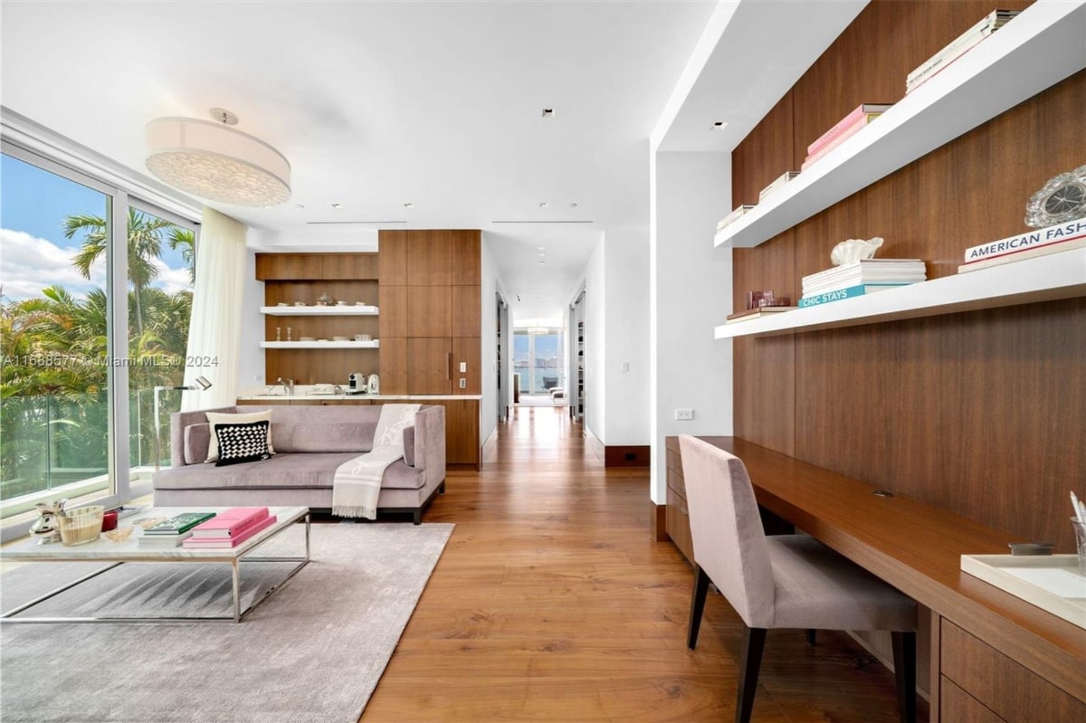 Living area with a light gray sectional sofa, a glass coffee table, and a built-in wooden desk and shelving unit. The floor is hardwood, and a light-colored rug is under the sofa. The space is open and bright, with large windows letting in natural light. A hallway leads further into the home.