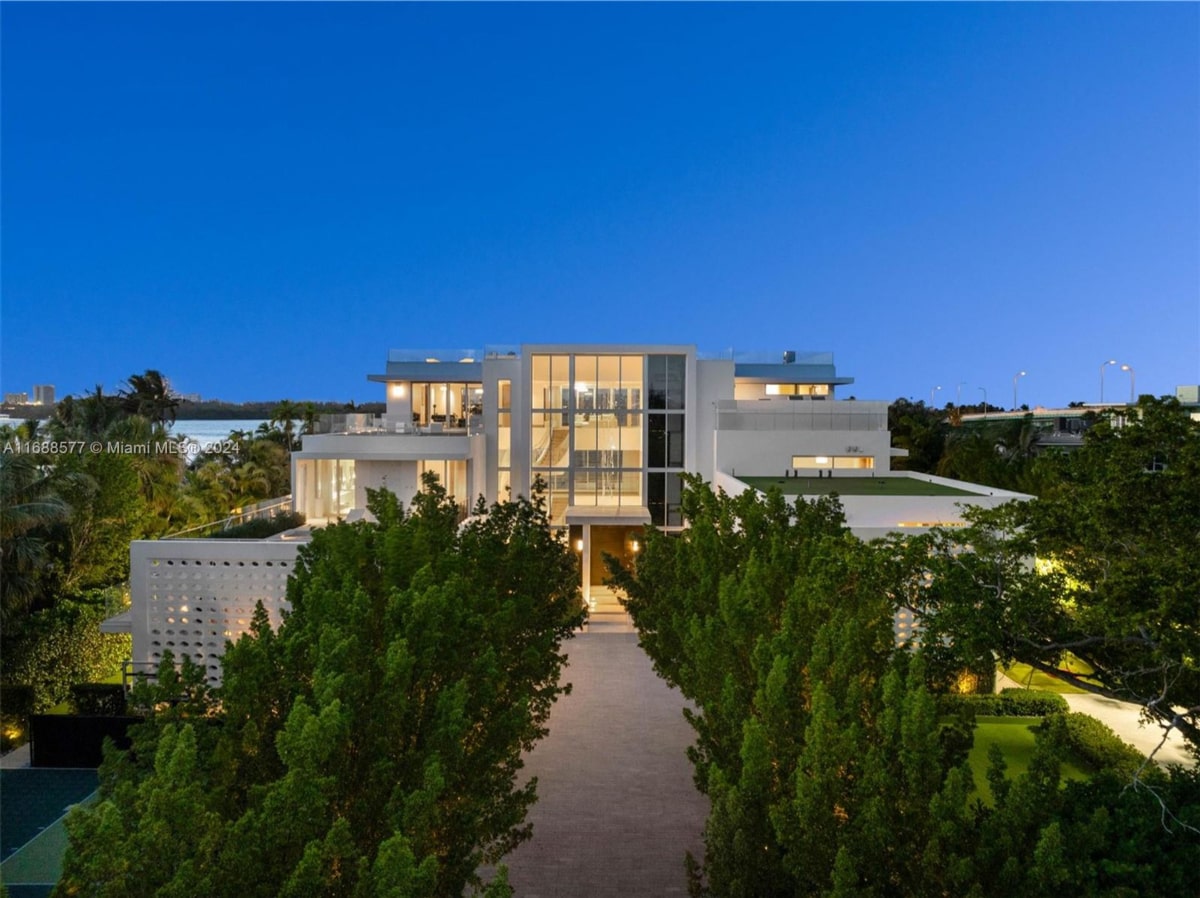 Evening view of a large, modern, white house. It has a flat roof and is set back from the road, with two rows of trees lining a long driveway leading to the entrance.