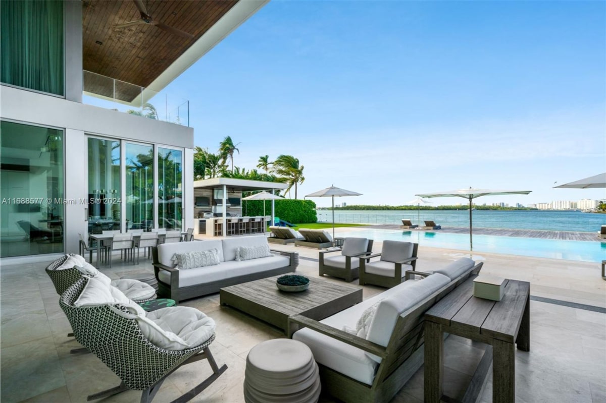 Outdoor living space overlooking a waterfront. There's a large sectional sofa, armchairs, and coffee tables arranged around a fire pit or similar feature. The area is spacious and elegantly furnished, with a large pool and a beautiful view of the water and distant shoreline in the background.