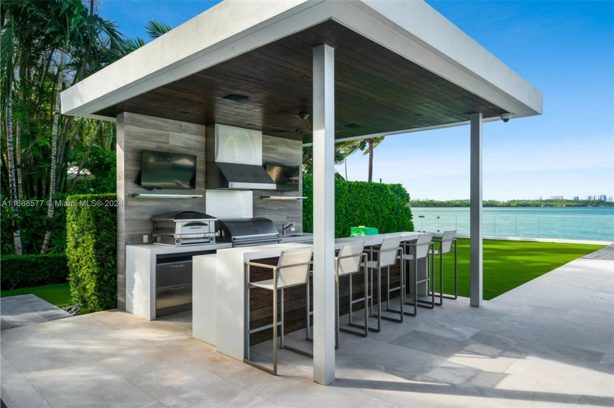 Outdoor kitchen with a covered pavilion. The kitchen is equipped with high-end appliances, including a grill and range hood. There's a bar area with stools, and the pavilion provides shade and protection from the elements. A view of water and greenery is visible in the background.