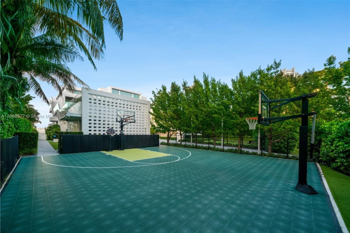 Private outdoor basketball court with a teal-colored surface. A basketball hoop is mounted on a pole at one end of the court. The court is surrounded by landscaping, including trees and some type of fence or wall.