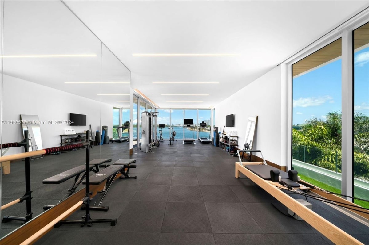 Home gym with floor-to-ceiling windows offering a view of the water. The gym contains various pieces of exercise equipment, including weight benches and a reformer Pilates machine. The flooring is dark gray or black.