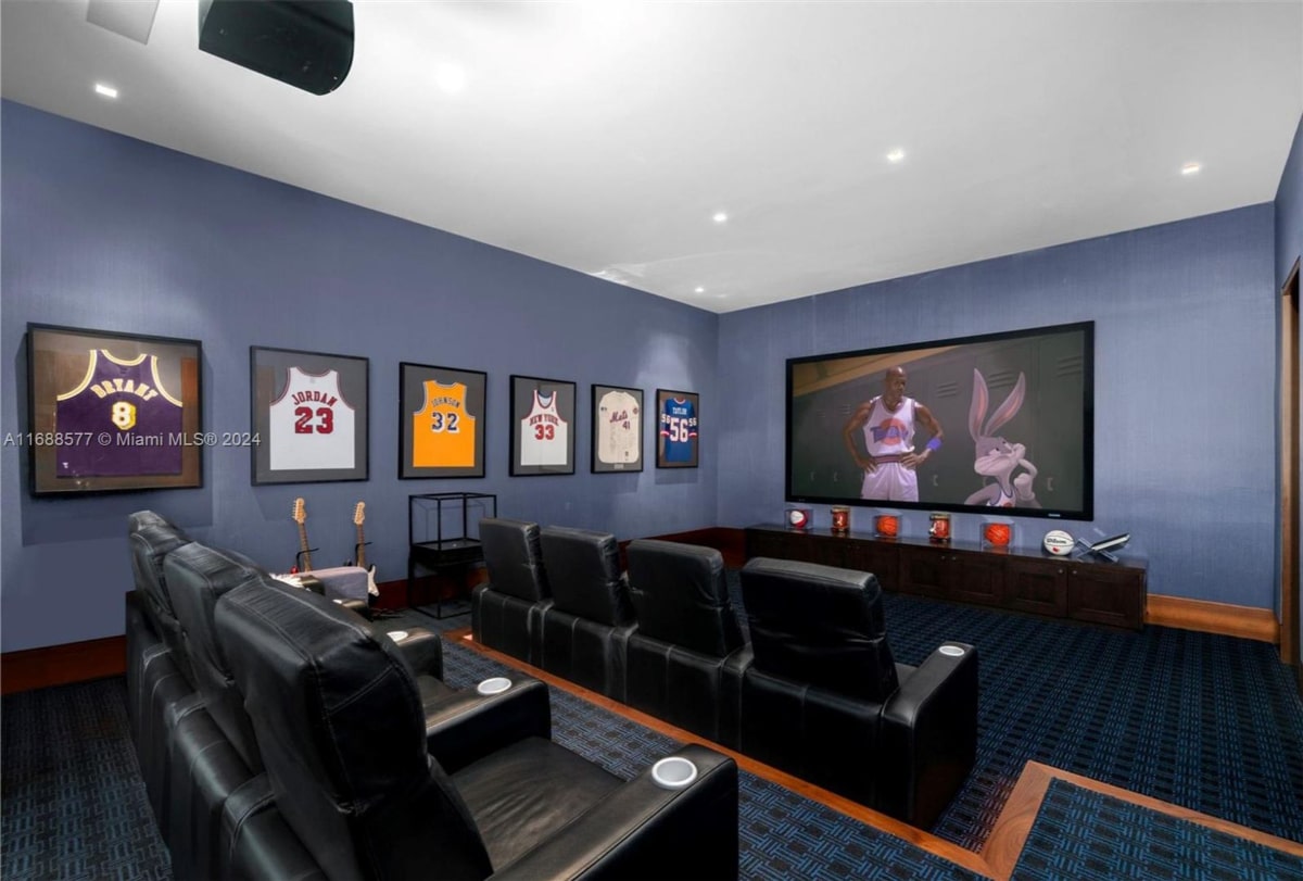 Home theater with a large screen displaying a scene from Space Jam. The room is painted a muted blue, and there are several black reclining chairs arranged for viewing. Framed basketball jerseys are displayed on the wall.