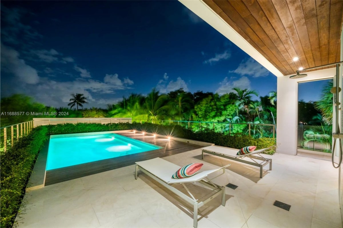 Nighttime view of a modern, minimalist pool area. The pool is rectangular and illuminated, with two lounge chairs positioned nearby. Lush greenery is visible beyond the pool area, and the overall atmosphere is serene and tranquil.