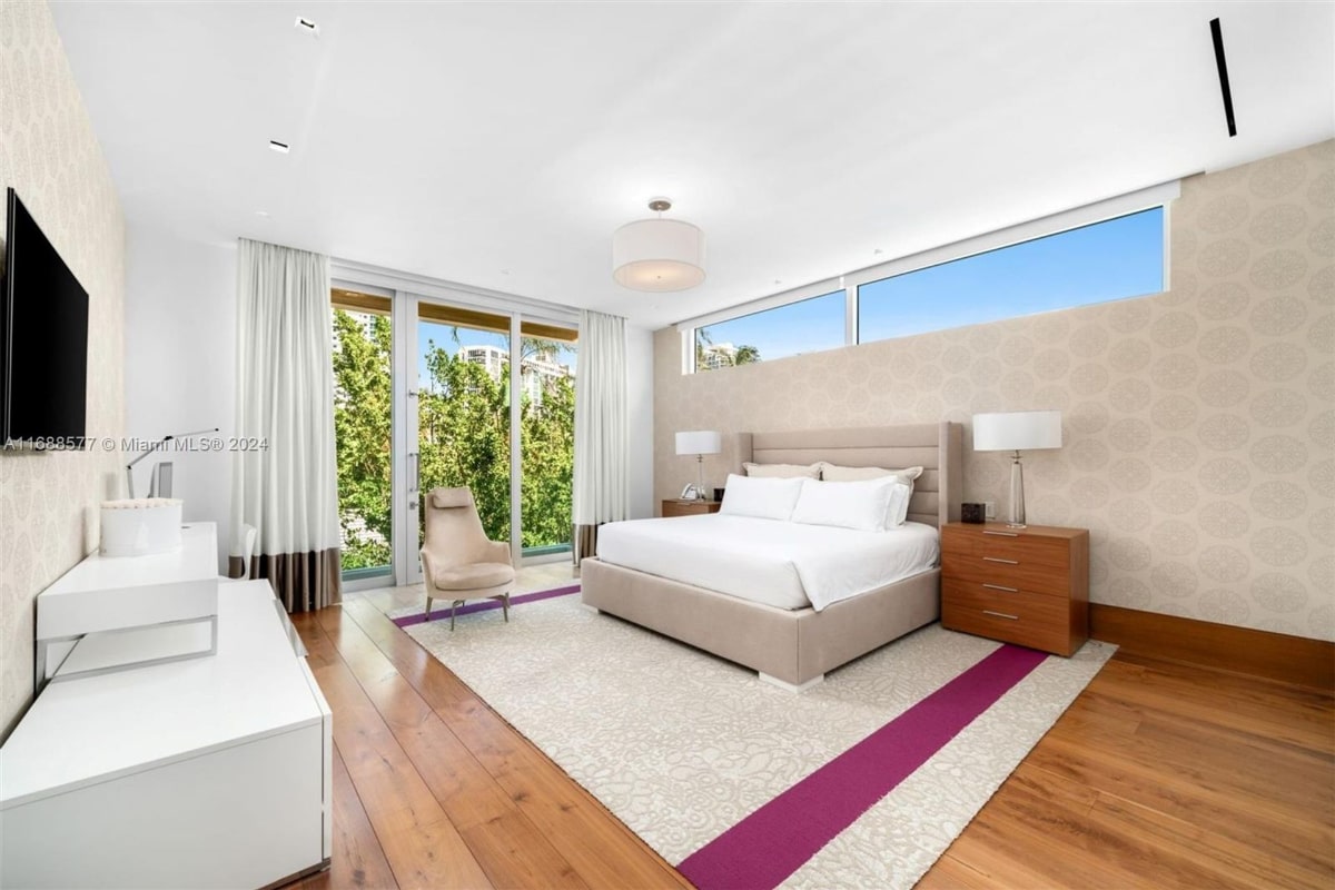 Master bedroom with a large, light-grey upholstered bed, white bedding, and two bedside tables. There are floor-to-ceiling windows and sheer curtains, allowing ample natural light. A light-colored armchair is placed near the windows, and a light-colored rug with a purple stripe is on the hardwood floor.