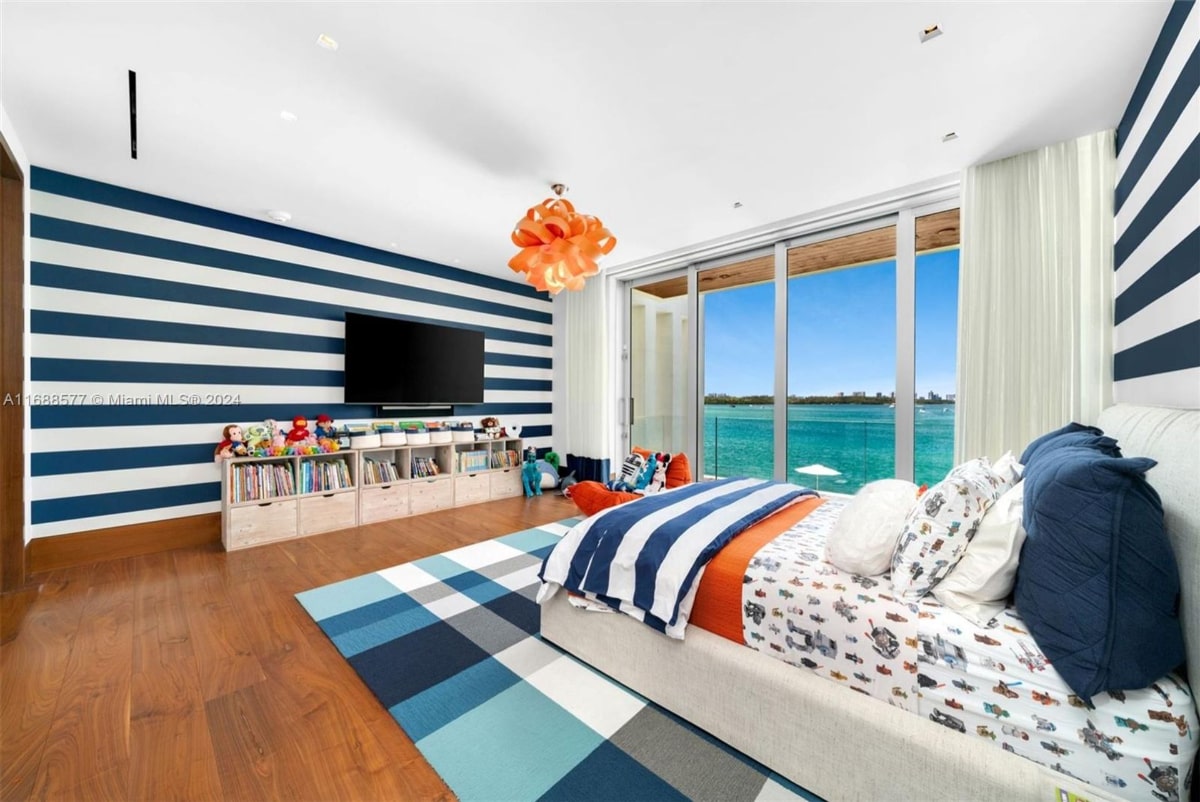  Child's bedroom with a nautical theme. The room features a bed with blue and white striped bedding, a navy and white striped wall, and a blue and gray plaid rug. There's a built-in storage unit, and floor-to-ceiling windows offer a stunning ocean view.