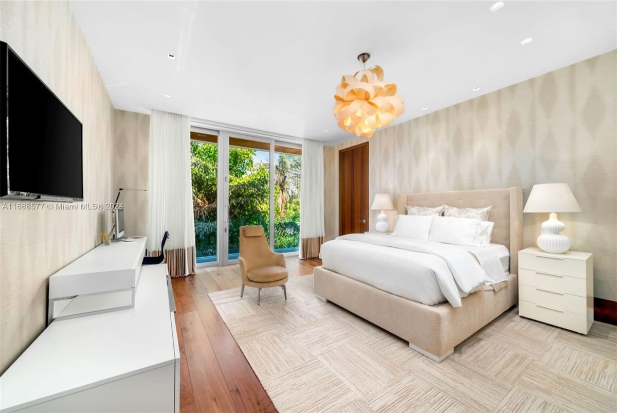 Bedroom with light beige upholstered bed, white bedding, and a light brown armchair. There are floor-to-ceiling windows with sheer curtains, offering a view of lush greenery. The floor is hardwood, and a light-colored rug is under the armchair. A modern light fixture hangs from the ceiling.