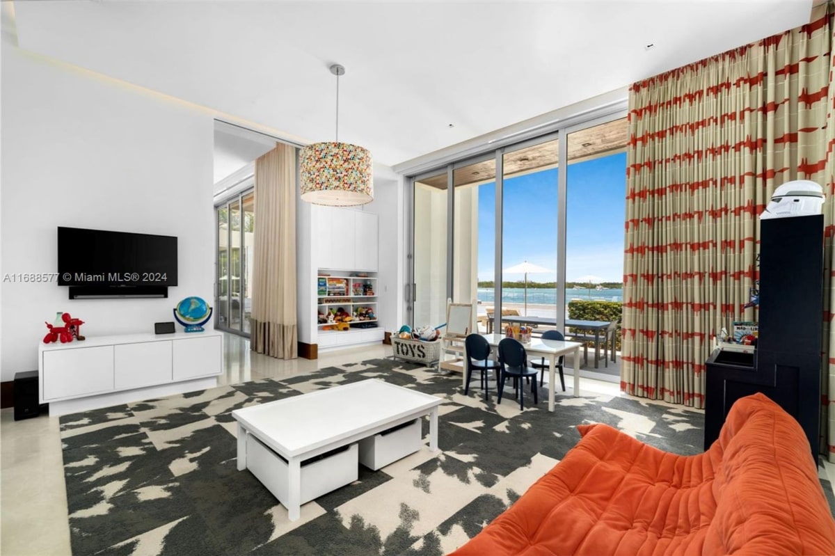 Bright and spacious playroom with floor-to-ceiling windows overlooking a body of water. There's a white coffee table, children's chairs and a table, a toy bin, and a coral-colored sofa or beanbag chair. The flooring is carpeted, and the overall style is modern and minimalist, designed for children's play.