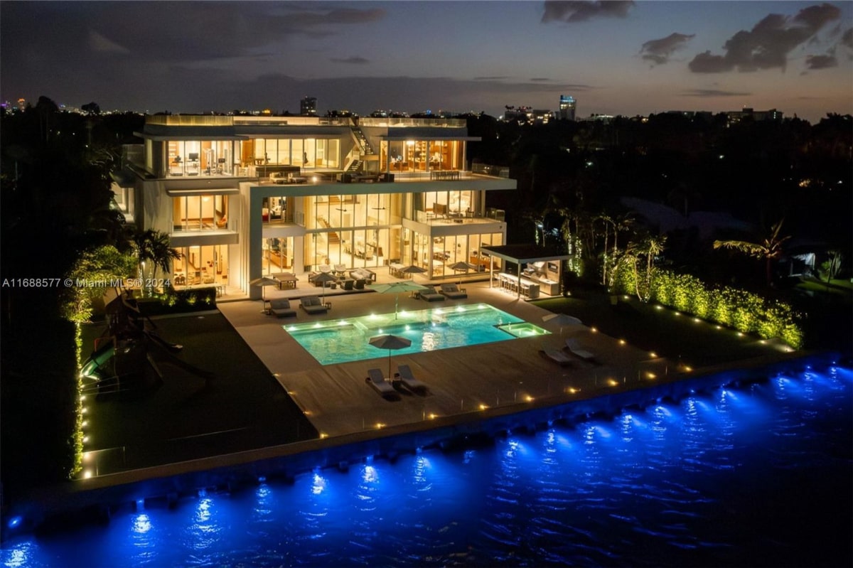 Aerial nighttime shot of a large, modern house with extensive outdoor lighting. The house is multi-storied with large windows, and it has a sizable rectangular pool illuminated from below. There is also what appears to be a secondary, smaller pool or spa adjacent to the main pool. The surrounding landscaping is dark, emphasizing the house and pool's lighting.