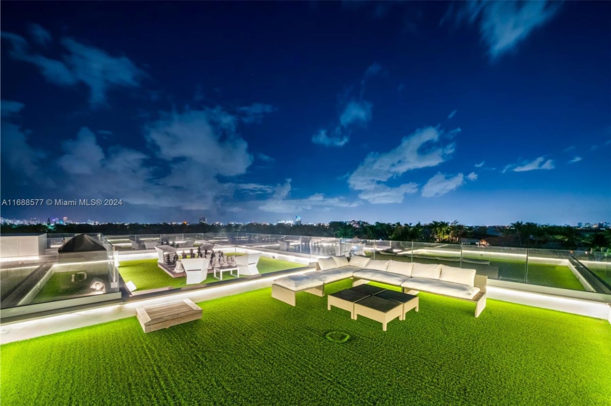 A rooftop terrace at night. The terrace features a large, white sectional sofa and coffee tables situated on artificial turf. There's also a low wooden bench or planter, and what looks like a bar area is visible in the background.